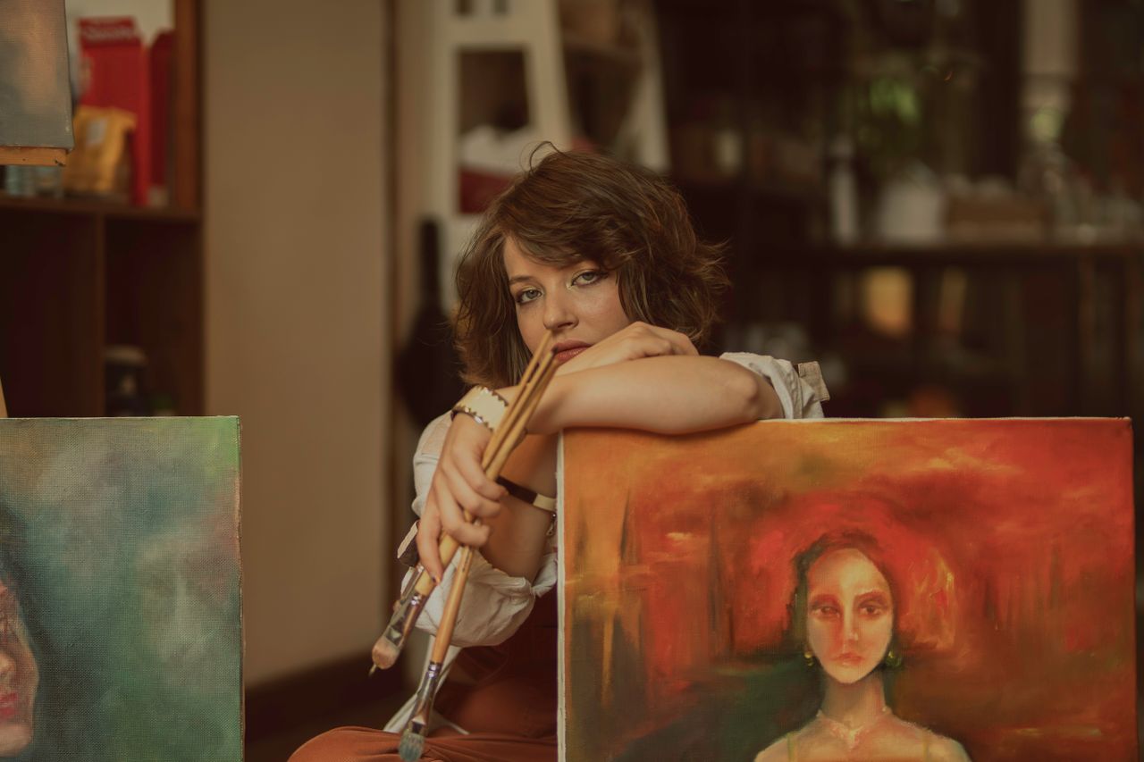 services-01 A woman artist in a studio with vibrant paintings, holding paintbrushes, showcasing creativity.