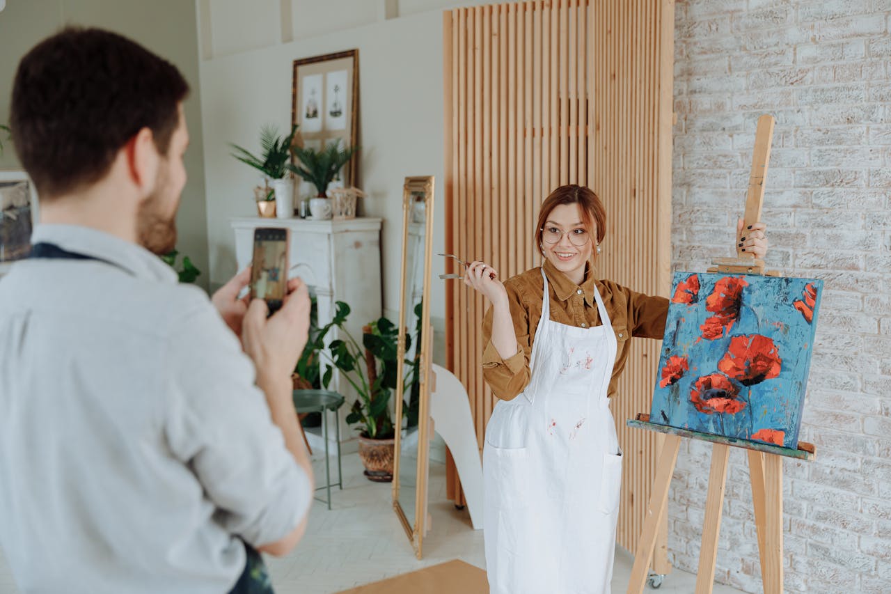 about-us-02 A woman artist proudly displays her painting while a man captures the moment on his phone.